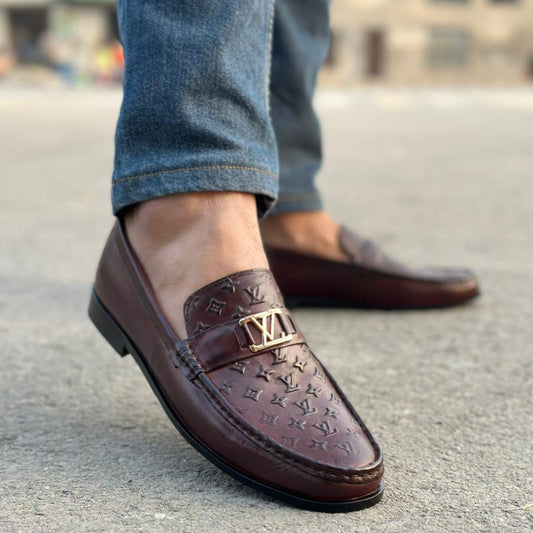 Brown loafers with a brand logo worn by a person on a concrete surface.